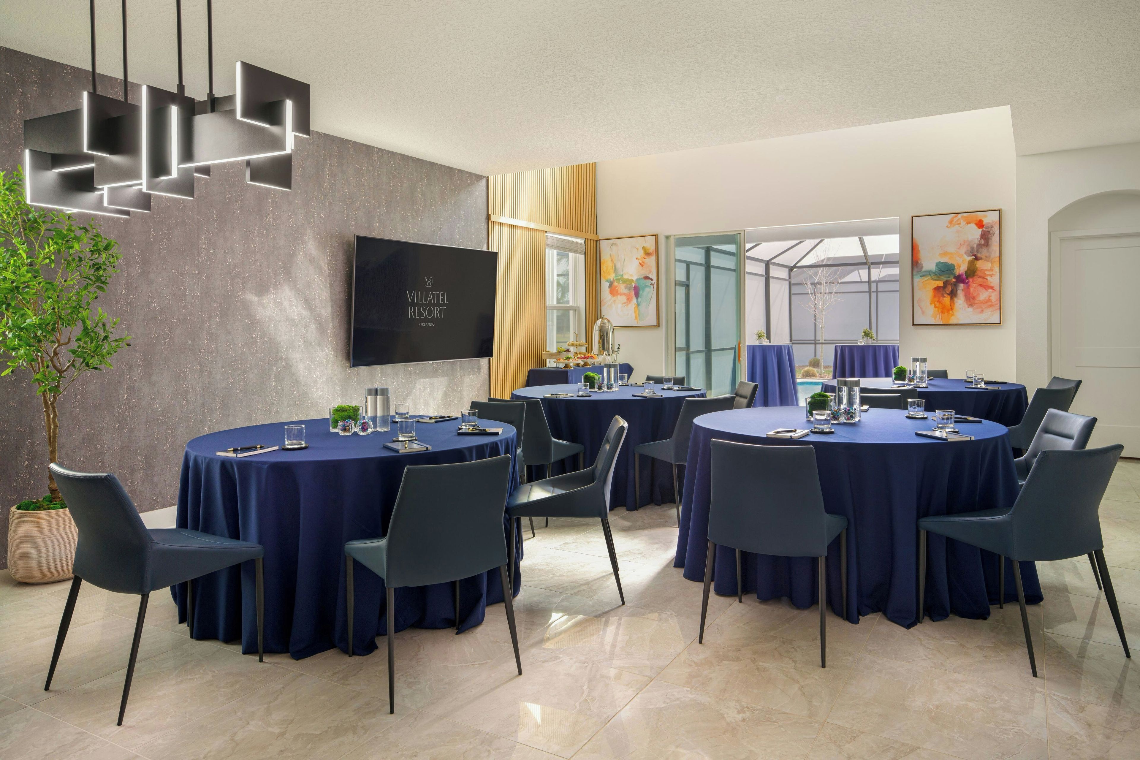 A conference room with round tables, chairs, a flat-screen TV, and a plant.
