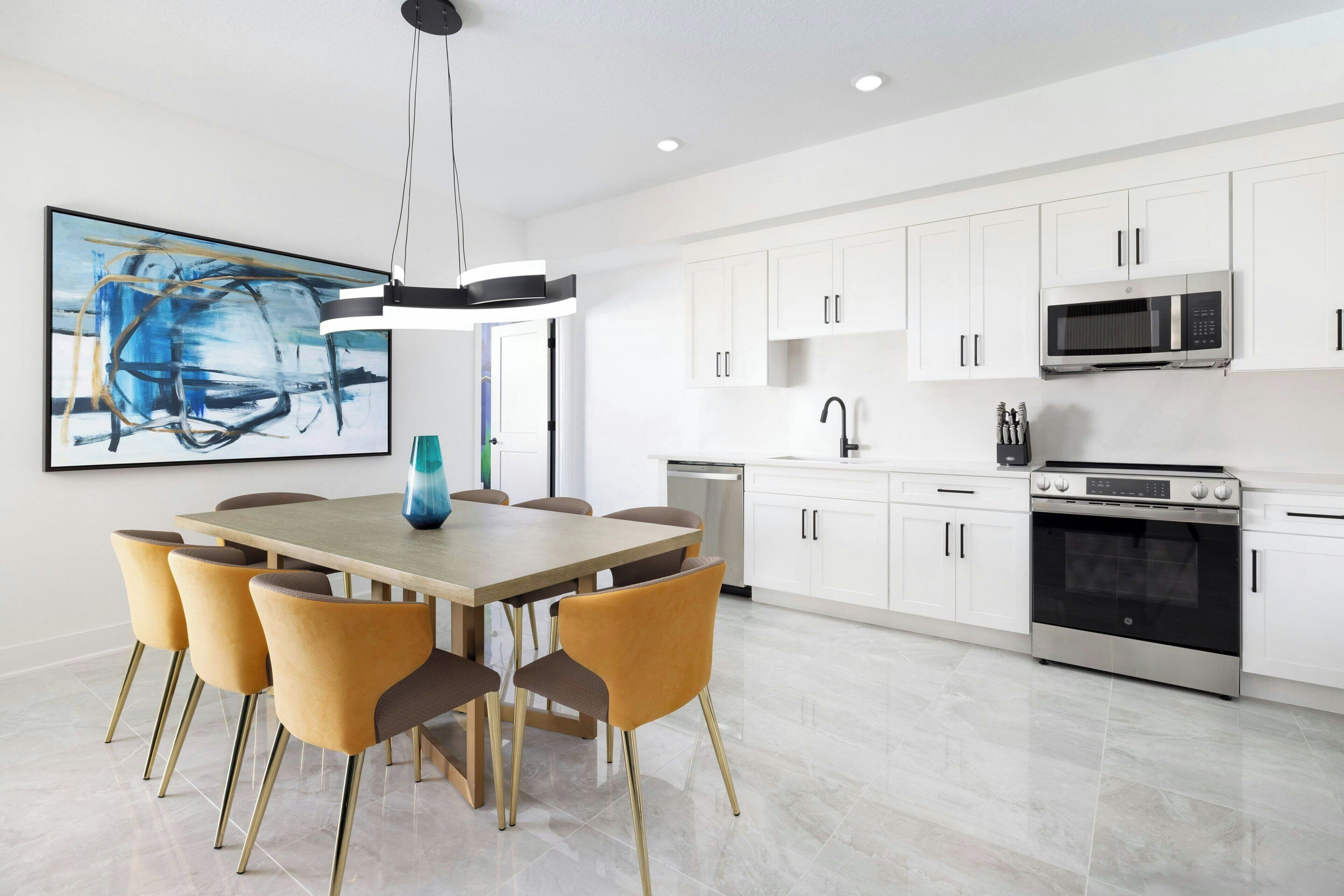 Una cocina moderna con armarios blancos, un microondas de acero inoxidable y un horno. Hay una mesa de comedor con sillas y un cuadro en la pared.