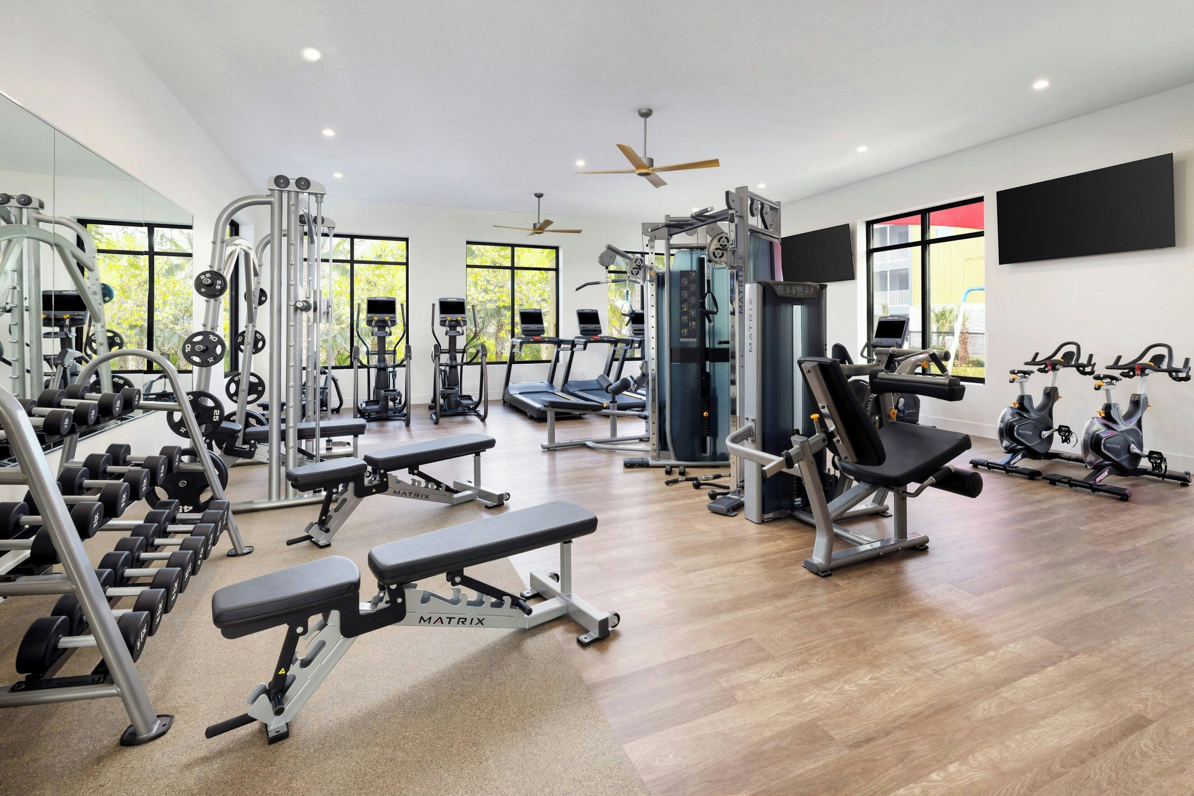 A modern gym with various exercise equipment, large windows, and ceiling fans.