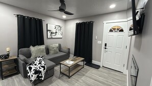 Een moderne woonkamer met een grijze bank, zwarte gordijnen, een plafondventilator, een lijstwerk en een witte deur.