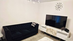 A living room with a black sofa, a white marble coffee table, a flat-screen TV on a white TV stand, and a modern chandelier.