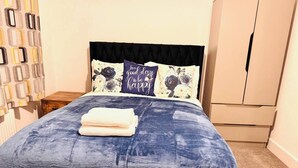 A neatly made bed with a blue bedspread and two pillows, one with floral patterns and the other with text. There is a wooden nightstand beside the bed, and a beige wardrobe in the background.