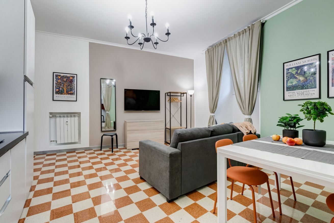 A modern living room with a checkered floor, a grey sofa, a dining table with orange chairs, a TV, and artwork on the walls.