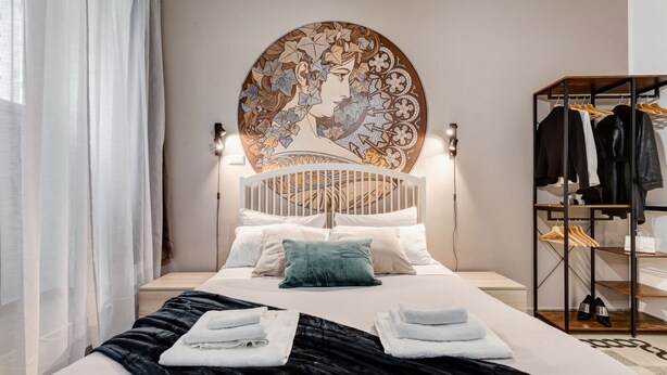 Habitación de hotel con una cama, cabecera con panel decorativo, armario y mesita de noche con lámpara.