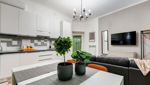 Una cocina moderna con armarios blancos, una mesa de comedor con dos plantas en macetas, un tapete gris y un sofá negro en una sala de estar con televisor.