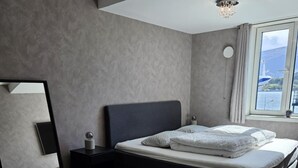 A modern bedroom with a large bed, a bedside table, a mirror, and a window with a view of the sea. - Free Parking Space, Balcony, Prime Location (Ålesund)