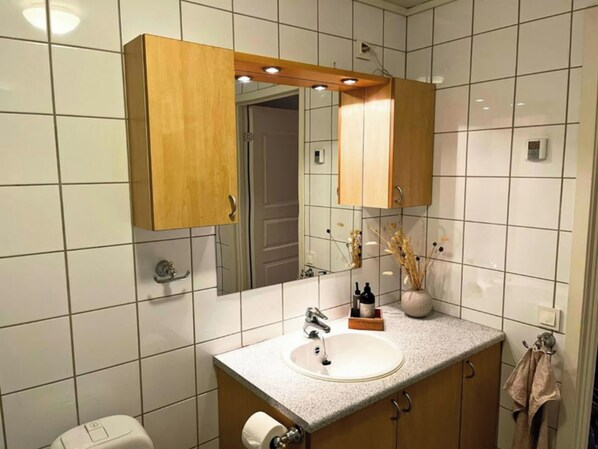 A bathroom with white tiled walls, a wooden vanity with a sink, a mirror, and a towel rack. - Free Parking Space, Balcony, Prime Location (Ålesund)