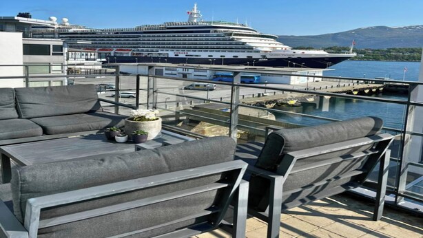 A balcony with grey sofas and a potted plant, overlooking a cruise ship docked at a port with mountains in the background. - Free Parking Space, Balcony, Prime Location (Ålesund)
