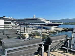 A balcony with grey sofas and a potted plant, overlooking a cruise ship docked at a port with mountains in the background. - Free Parking Space, Balcony, Prime Location (Ålesund)