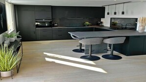 A modern kitchen with dark cabinetry, a central island with bar stools, and a large window providing natural light. - Free Parking Space, Balcony, Prime Location (Ålesund)