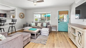 A modern living room with a sofa, coffee table, and chairs. There is a door with a window, a ceiling fan, and a cabinet with a TV. - Coastal Vibes Beach House (Oceanside)