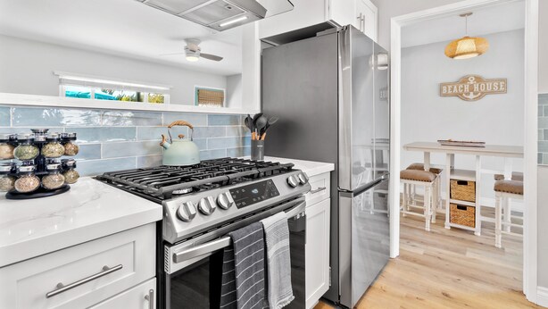 A modern kitchen with a stainless steel refrigerator, a gas stove, and a built-in oven. There is a small island with stools and a sign that reads "BEACH HOUSE". - Coastal Vibes Beach House (Oceanside)