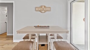 A dining area with a table and benches, a wall-mounted sign reading "BEACH HOUSE", and a view through a glass door. - Coastal Vibes Beach House (Oceanside)