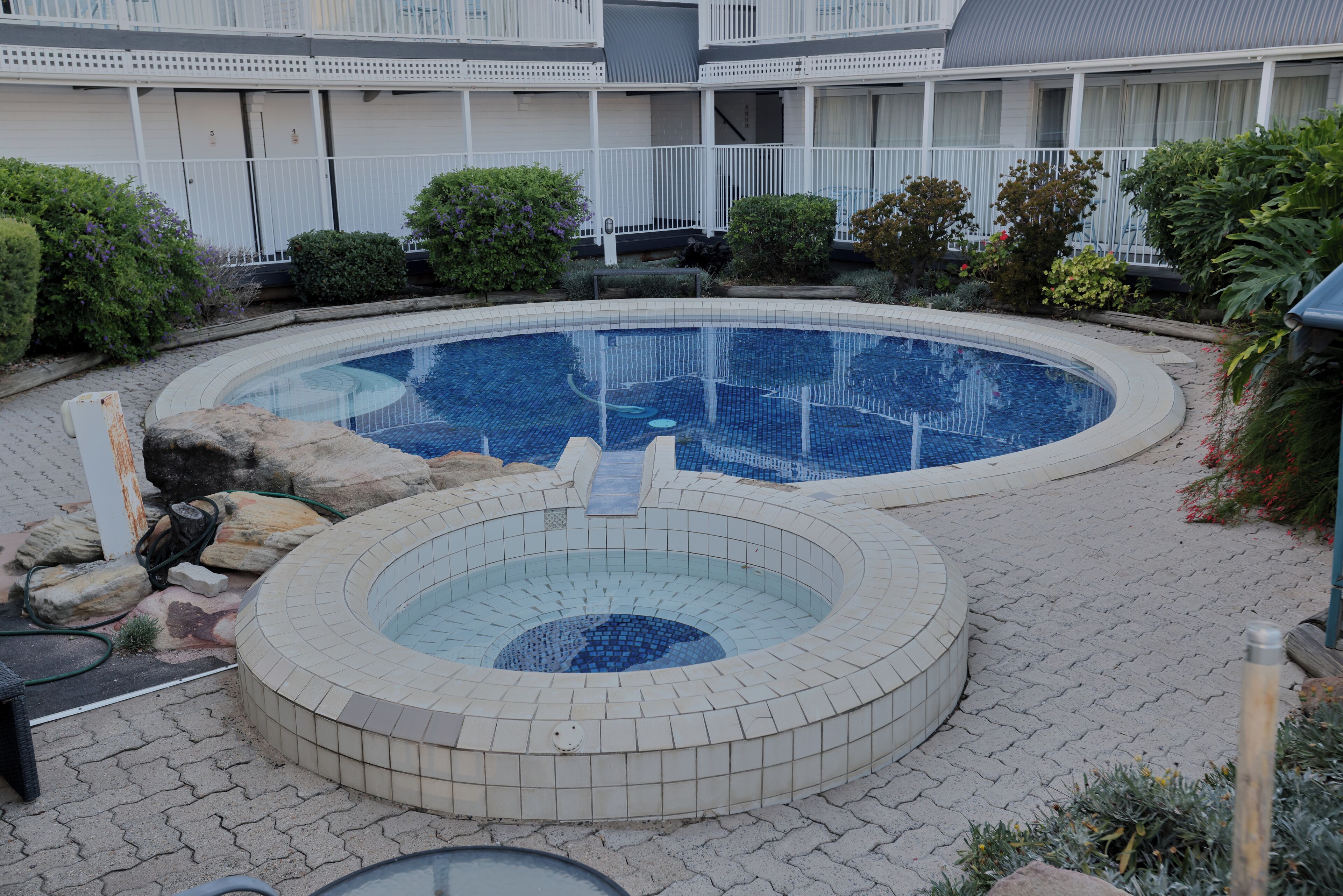 A circular pool with a hot tub in the center, surrounded by a paved patio area with a white fence and greenery.