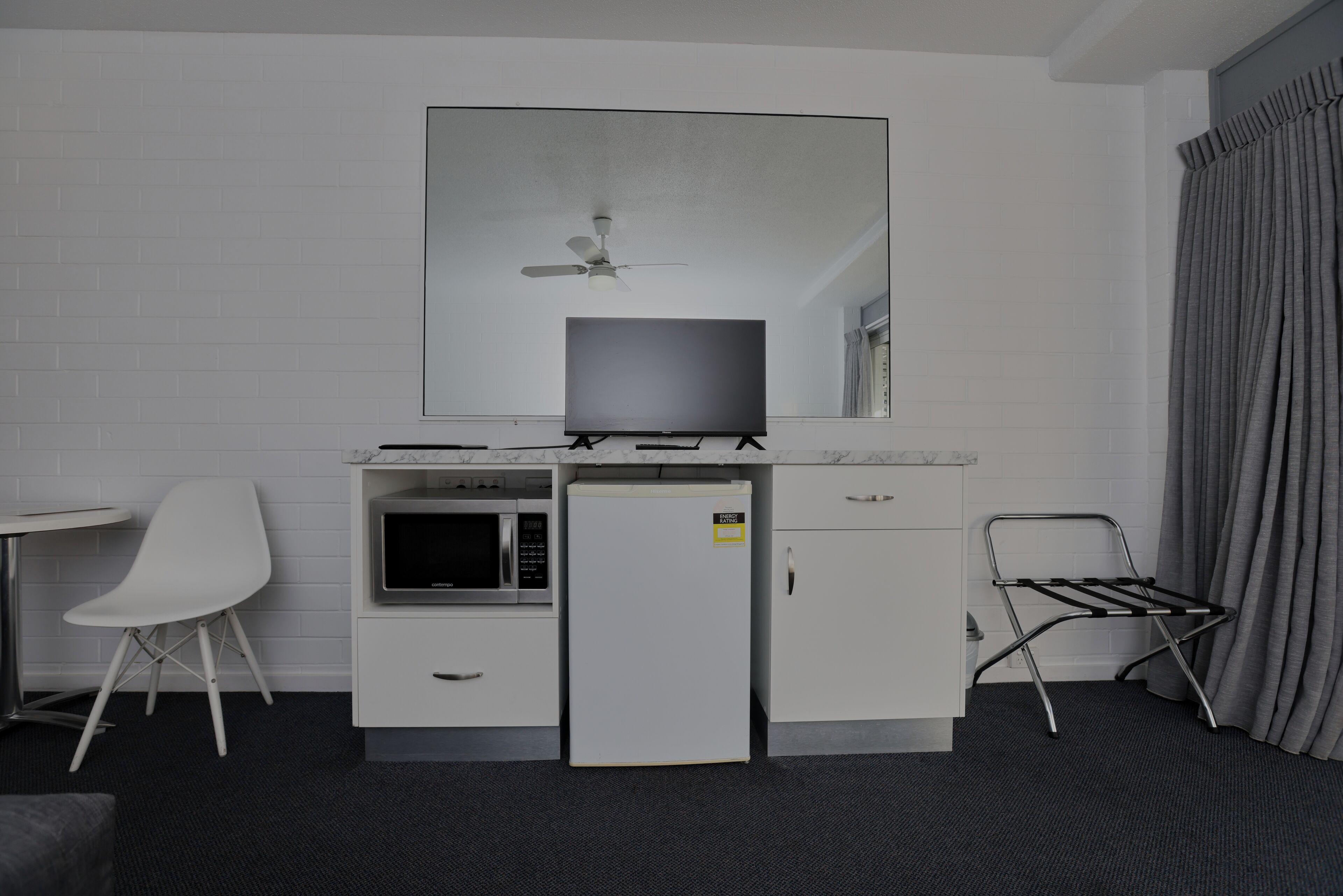 A compact hotel room with a kitchenette, a flat-screen TV, a ceiling fan, a white chair, a small refrigerator, and a drying rack.