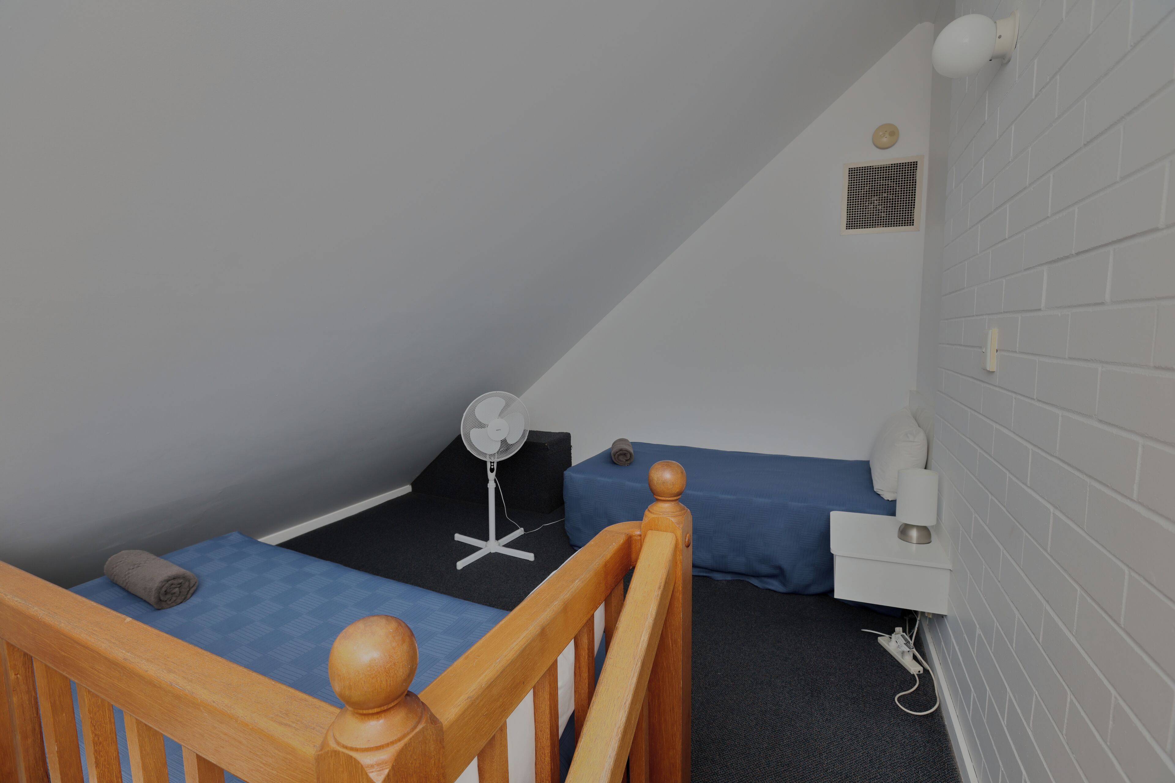 Un dormitorio pequeño y con una sola habitación, con estructura de cama de madera, un escritorio, una silla, un ventilador y una pared blanca con una rejilla de ventilación.