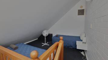 Un dormitorio pequeño y con una sola habitación, con estructura de cama de madera, un escritorio, una silla, un ventilador y una pared blanca con una rejilla de ventilación.