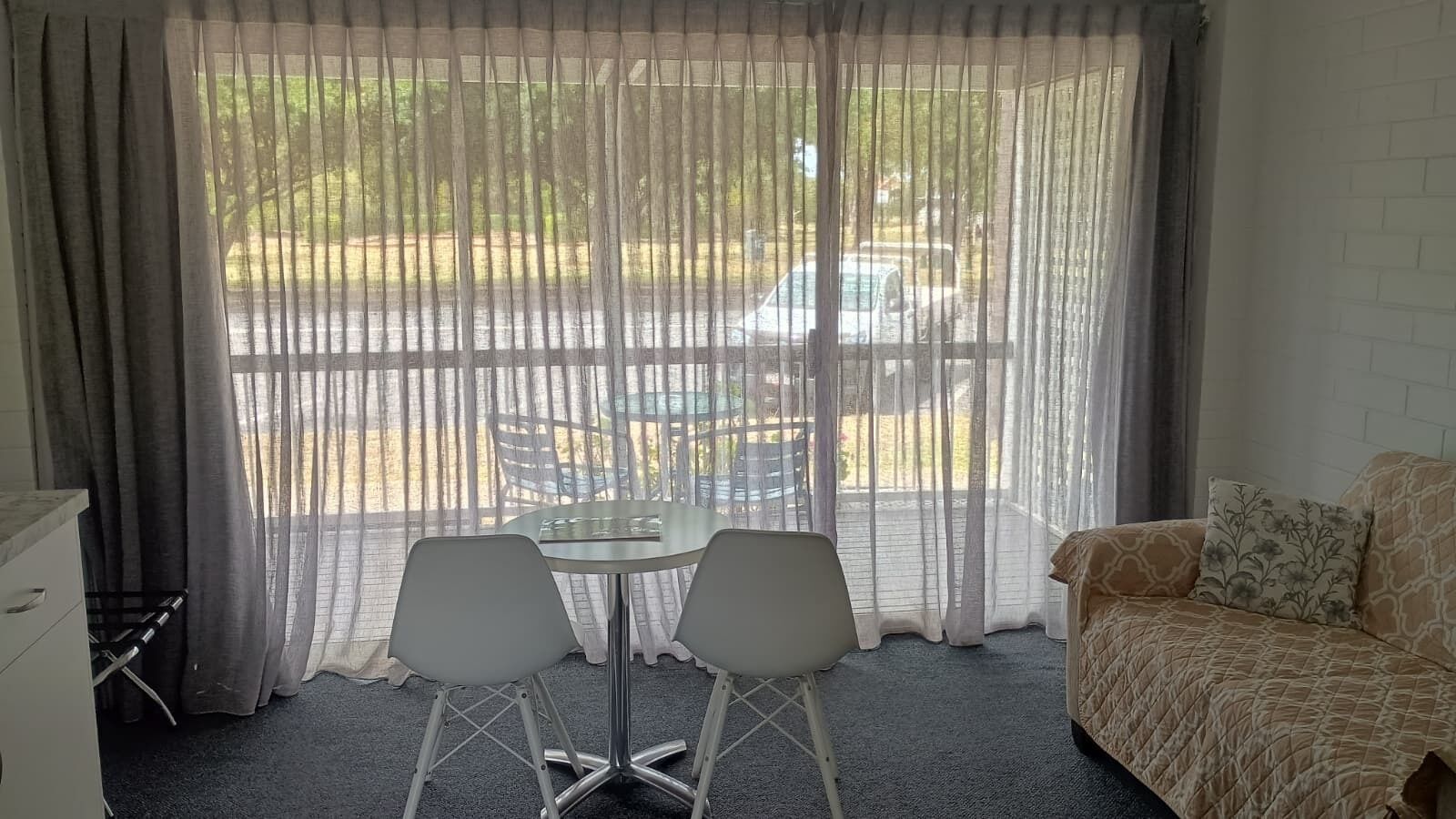 Ein Zimmer mit einem Bett, einem Tisch mit zwei Stühlen und einem Fenster mit durchsichtigen Vorhängen.