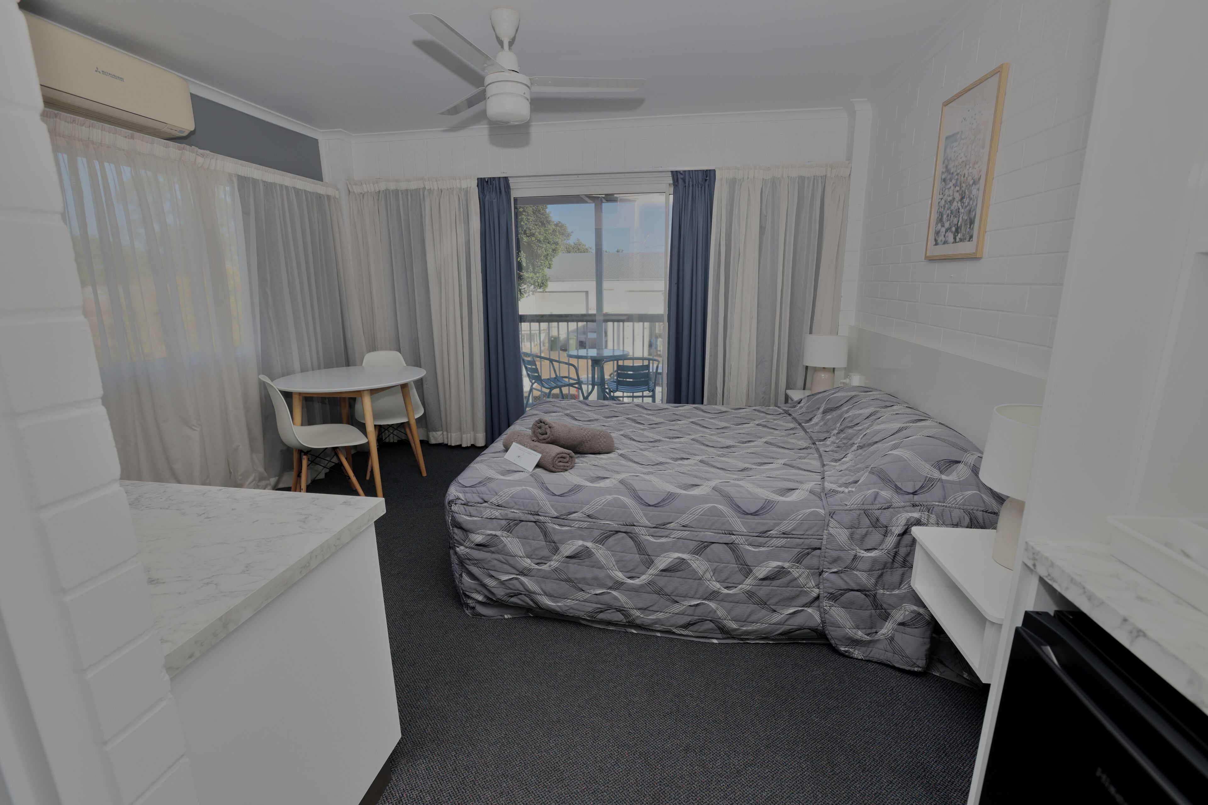 A compact hotel room with a bed, a small desk, a chair, a ceiling fan, and a window with curtains.