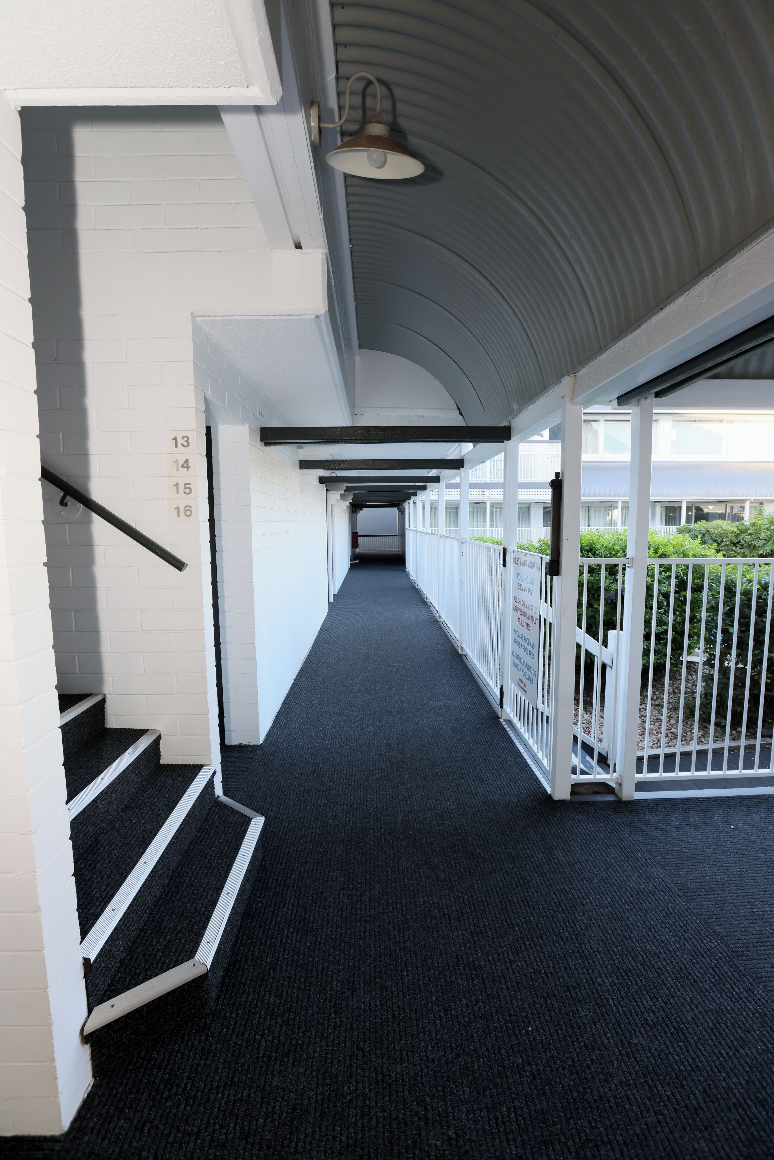 Un couloir avec un escalier, des murs blancs et une rambarde en métal.