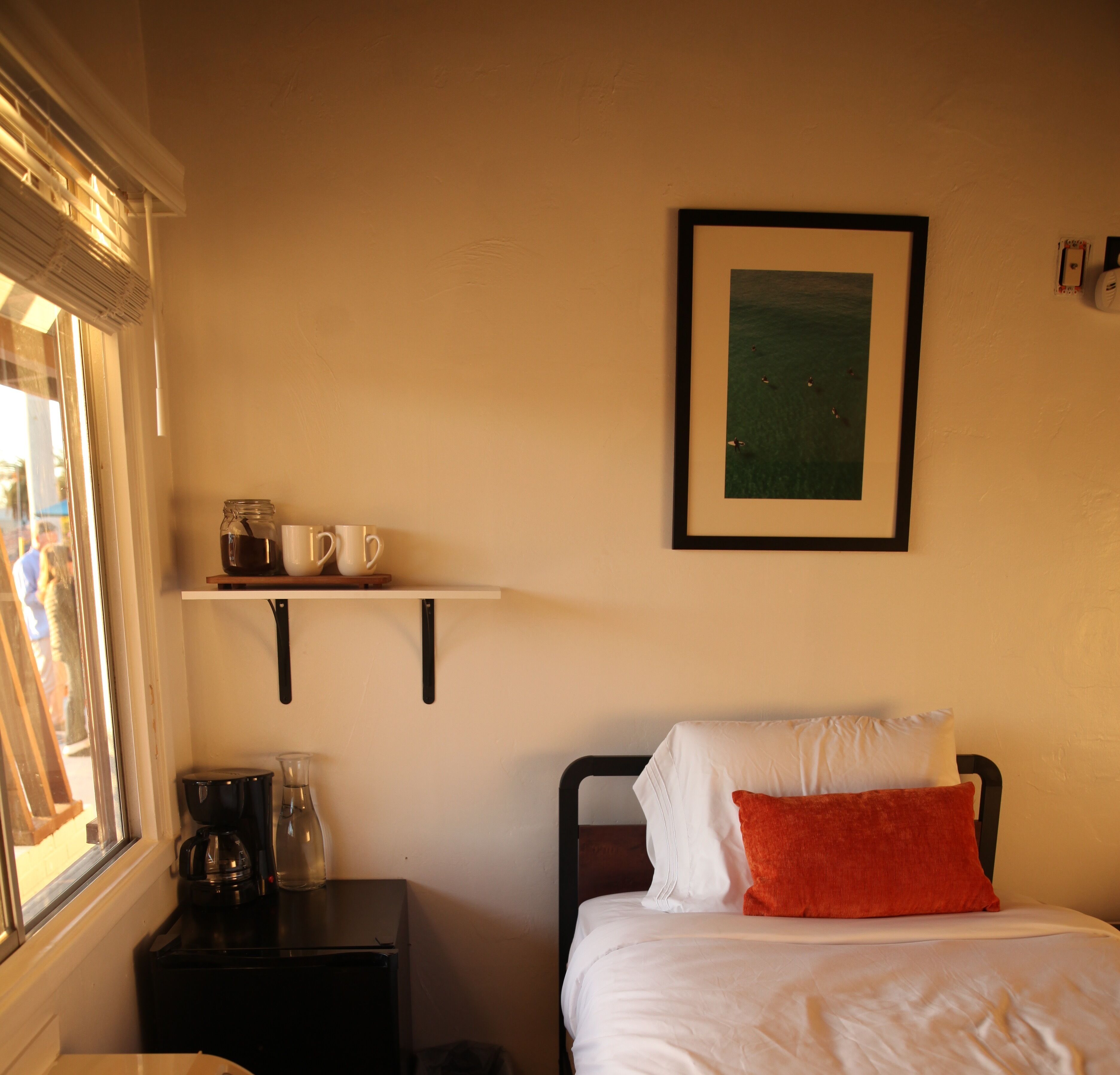 A bedroom with a bed, a nightstand, a coffee maker, a glass pitcher, and a shelf with mugs.