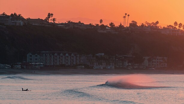 A coastal scene with a sunset, buildings, and a person surfing. - West Coast Surf and Stay (San Clemente)