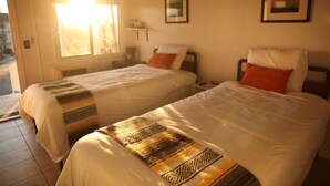 Two beds with white bedding and patterned blankets, a nightstand with a lamp, and a window with sunlight streaming in. - West Coast Surf and Stay (San Clemente)