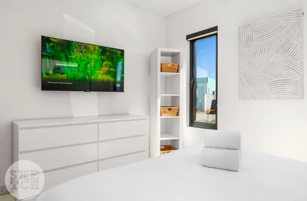 Un dormitorio moderno con televisión de pantalla plana montada en la pared, un armario blanco, una estantería empotrada y un amplio ventanal con vistas al exterior.