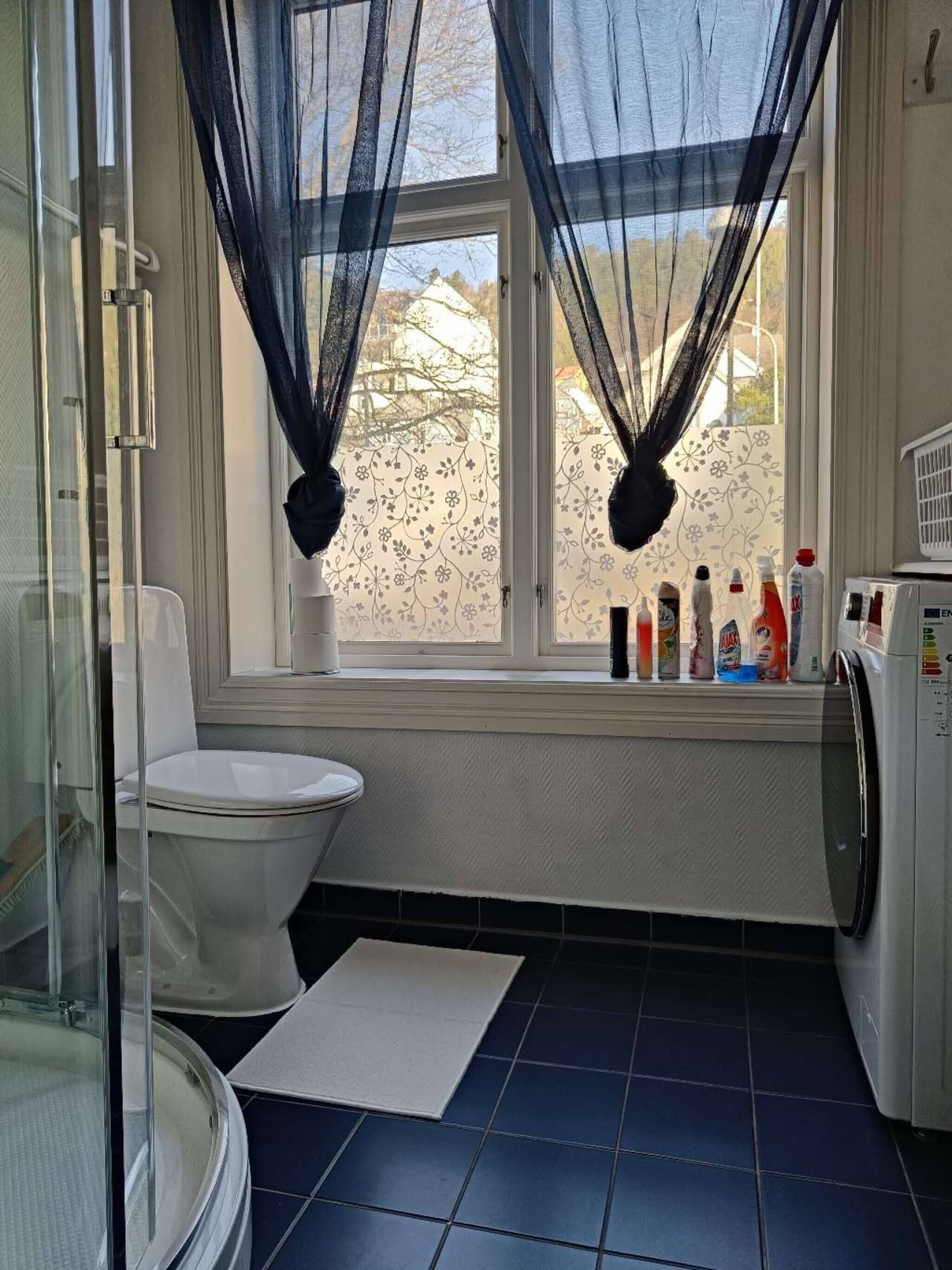 Egy fürdőszoba, amelyben található egy WC, egy üvegfalas zuhanyzó, egy mosógép és egy ablak, melyen könnyű, áttetsző függöny van.