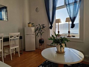 Een kleine, gezellige kamer met een ronde tafel, twee stoelen, een koelkast, een potplant en een raam met gordijnen.