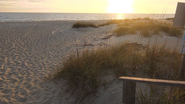 A beach with sand and grass, a wooden structure, and a sunset in the background.