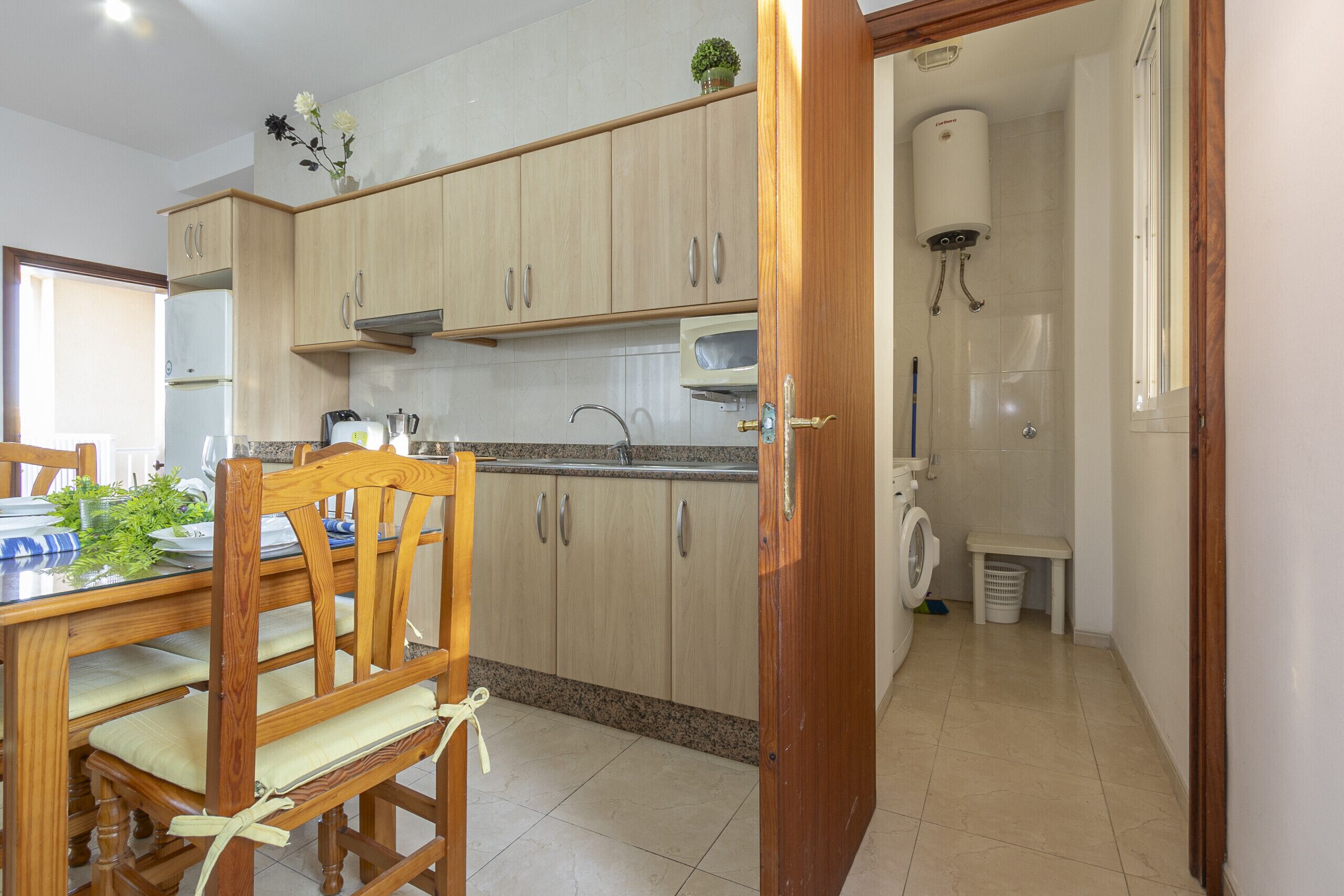 Een keuken met houten kastjes, een eettafel met stoelen, een ingebouwde magnetron en een boiler.