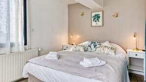 A neatly made bed with a quilted blanket, two bedside lamps, a framed plant, and a radiator.