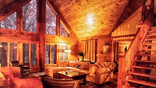 A cozy log cabin interior with a wooden staircase, large windows, and a fireplace.
