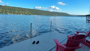 A wooden deck with red Adirondack chairs and a pair of shoes, overlooking a calm lake with a forested shoreline.