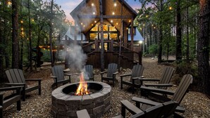 Una cabaña con un hogar de piedra rodeado de sillas Adirondack, ubicada en un bosque con luces colgantes.