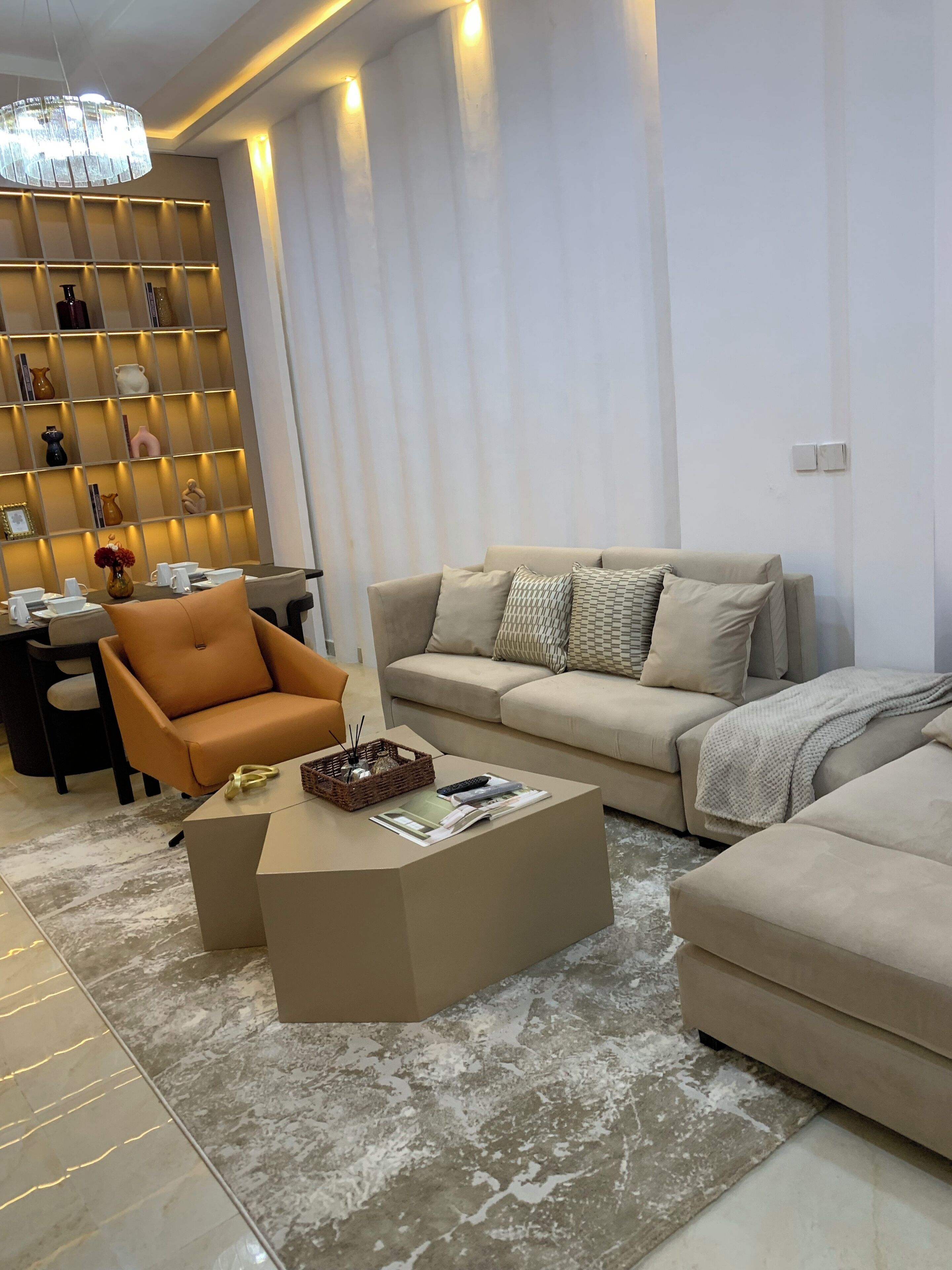 A modern living room with a beige sofa, a brown armchair, and a coffee table. There is a bookshelf with decorative items and a chandelier.