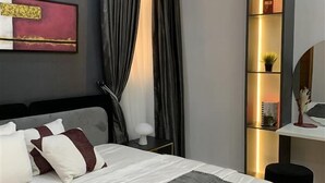 A modern bedroom with a bed, a bedside table, a desk with a chair, a wall-mounted air conditioner, and a shelf with decorative items. - Spacious 2-bedroom apartment (Lekki)