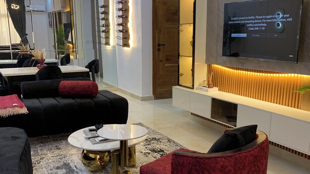 A modern living room with a large flat-screen TV mounted on the wall, a black sofa, a red armchair, and a round coffee table. - Spacious 2-bedroom apartment (Lekki)