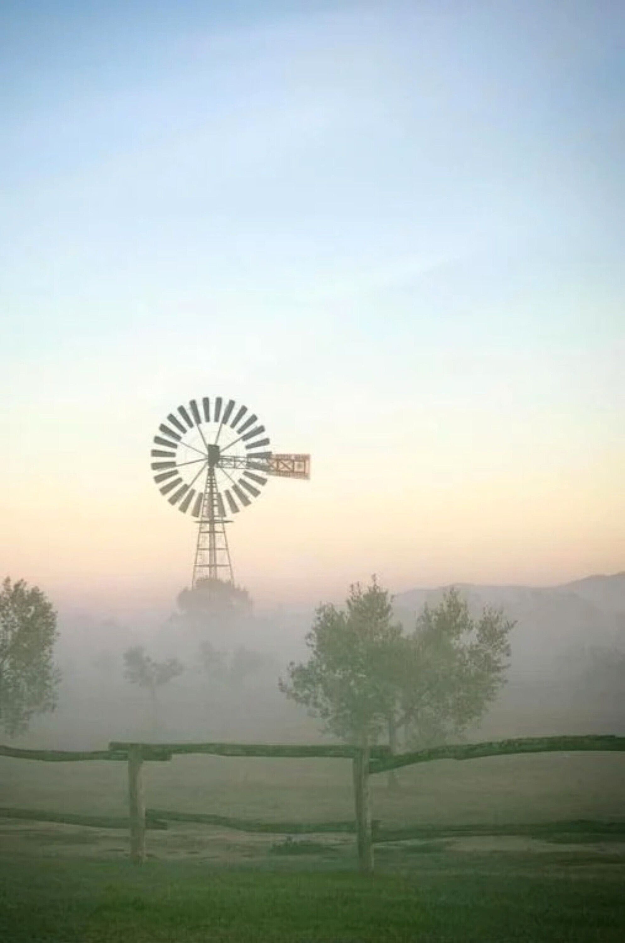 A windmill in a foggy landscape with a fence in the foreground.