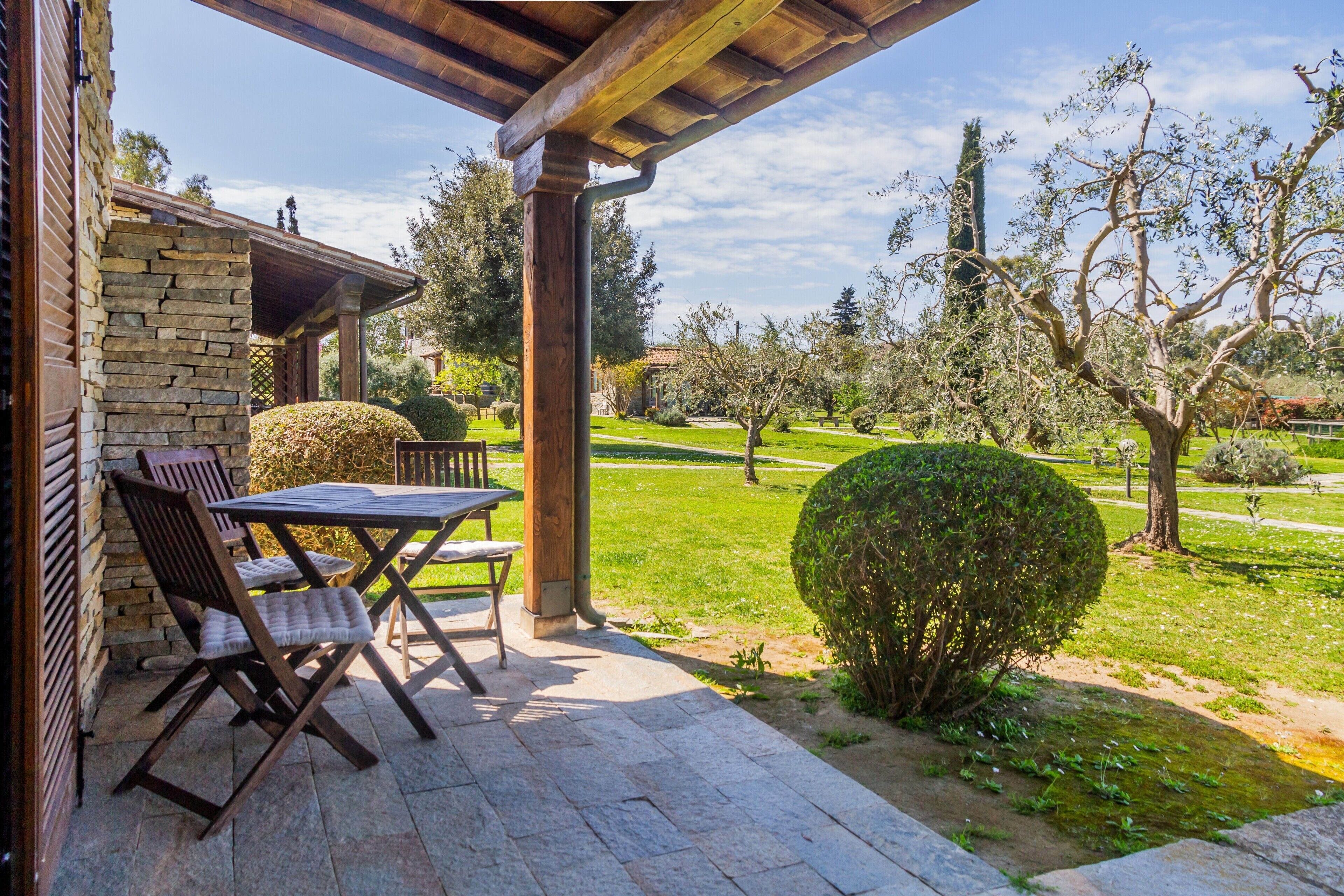 A patio with a stone table and chairs, a stone wall, and a view of a well-maintained garden with trees and a hedge.