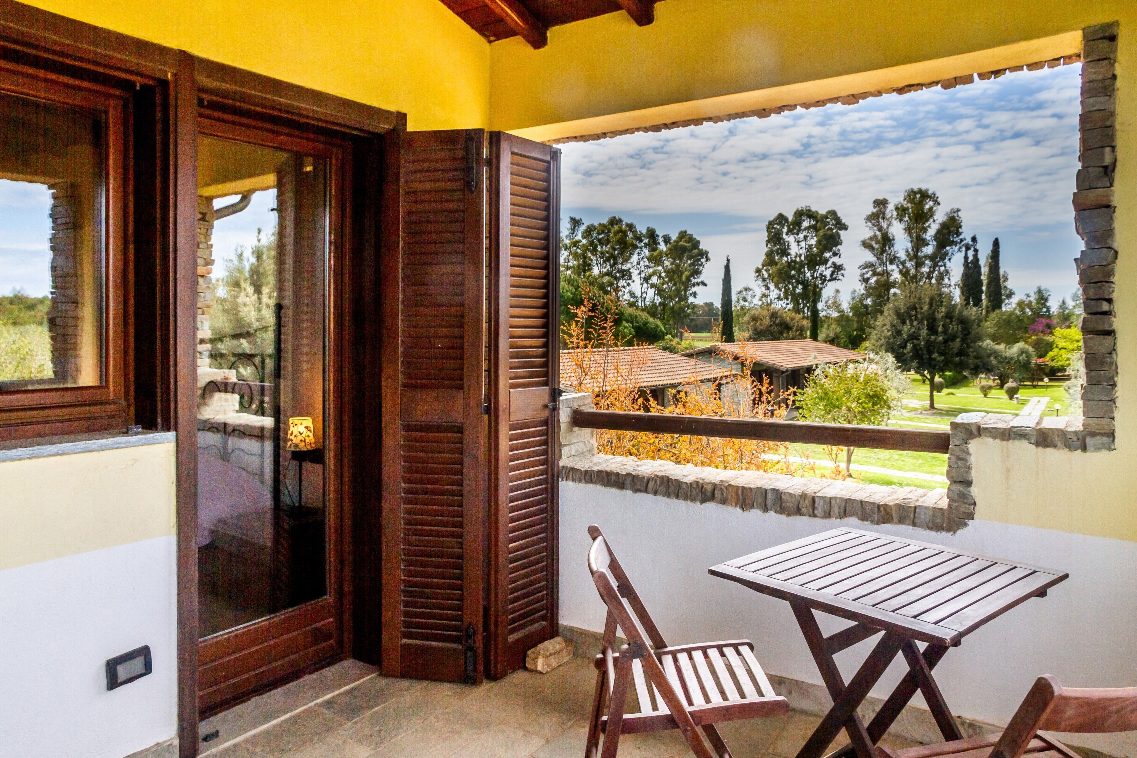 A balcony with a table and chairs, a wooden door, and a view of a garden and trees.