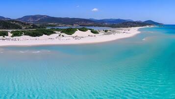 Un paysage côtier avec une plage de sable, des eaux turquoise et limpides, et des montagnes à l’horizon.