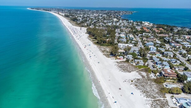 Luftaufnahme einer Küstenregion mit einem langen Sandstrand, angrenzenden Wohngebäuden und einem klaren, blauen Ozean.