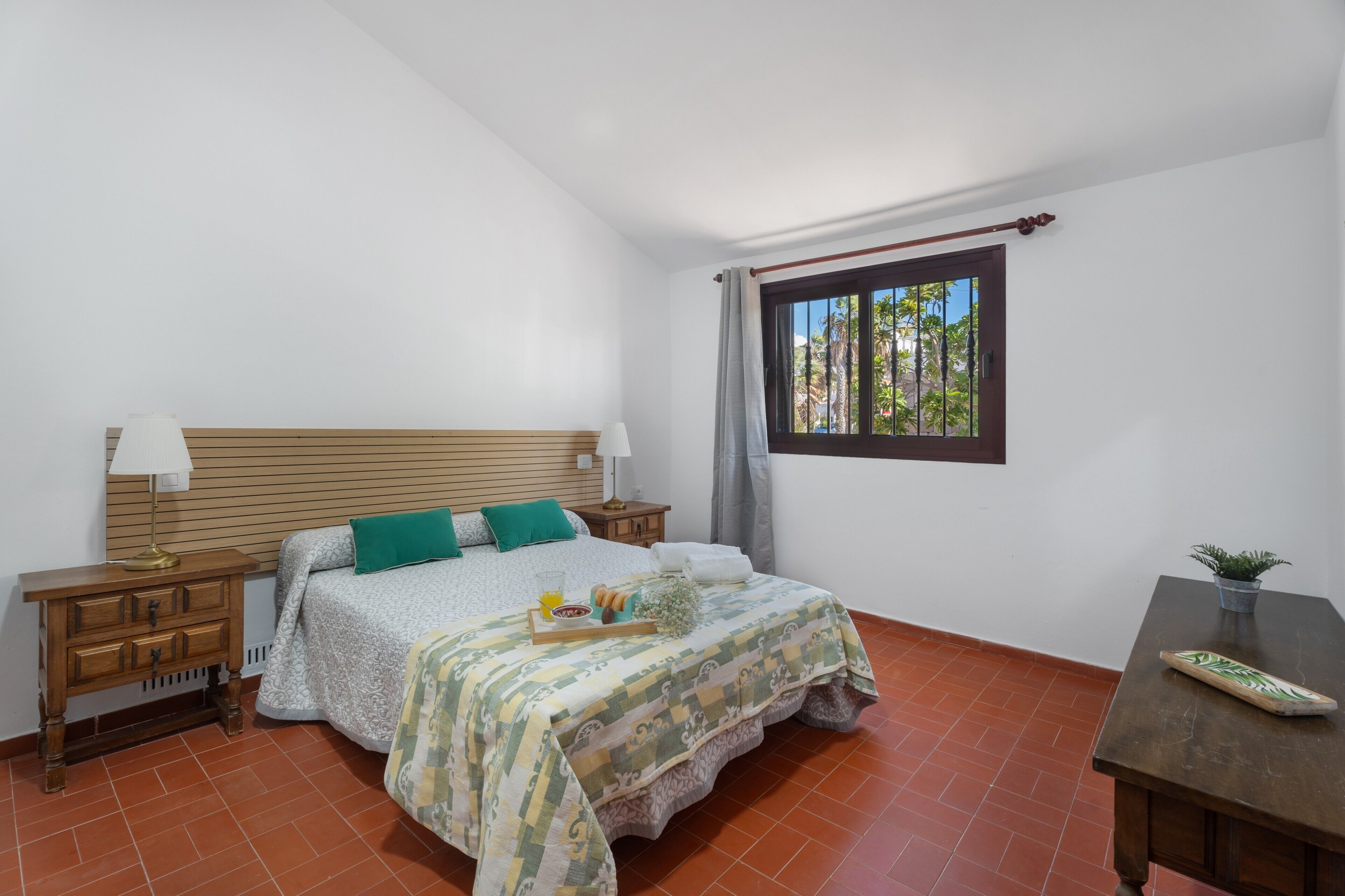 Une chambre à coucher avec un lit, des tables de chevet en bois, une fenêtre donnant sur un beau paysage, une petite table et une plante en pot.
