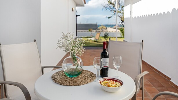 Mesa redonda con una botella de vino, dos copas, un bol de comida y un florero con flores en un balcón.