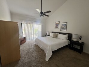 Un dormitorio con cama, mesita de noche, lámpara, ventilador de techo y ventana con persianas.