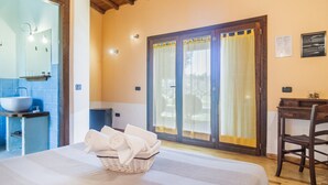 Ein Schlafzimmer mit Bett, Nachttisch, Stuhl, einem Fenster mit Vorhängen und einem Badezimmer mit Waschbecken und Spiegel.
