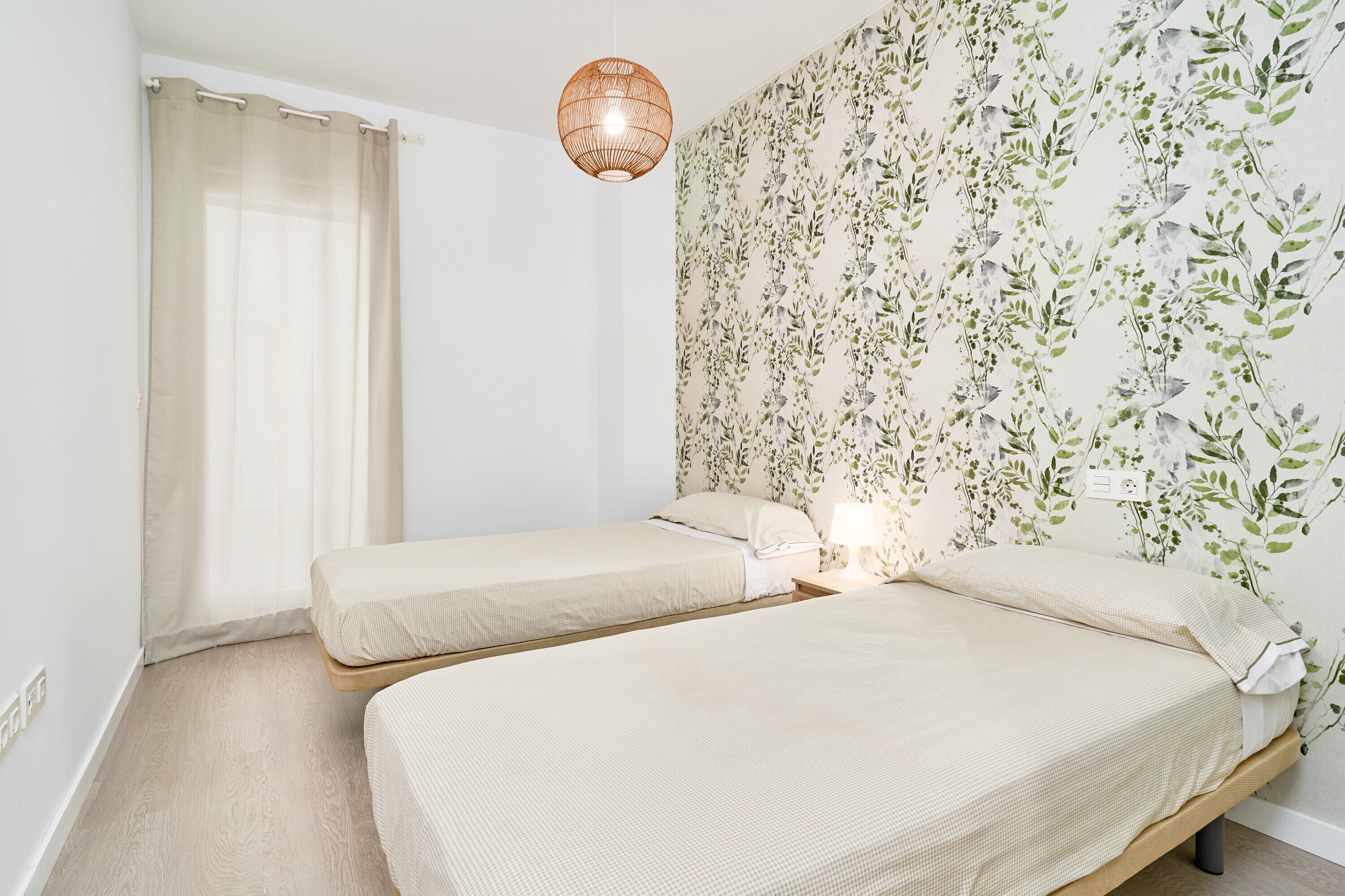 Quarto com duas camas, chão de madeira, parede com papel de parede floral, luminária pendente e cortina.