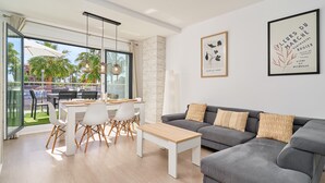 Una sala de estar moderna con un sofá gris, una mesa de centro de madera y un comedor con sillas blancas y una mesa de vidrio.